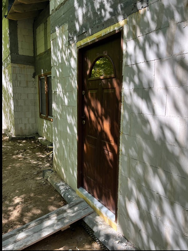 Ściana budynku z bloczków, okno i brązowe drzwi wejściowe z półokrągłym przeszkleniem, zabezpieczone żółtą taśmą. Widoczne cienie drzew.