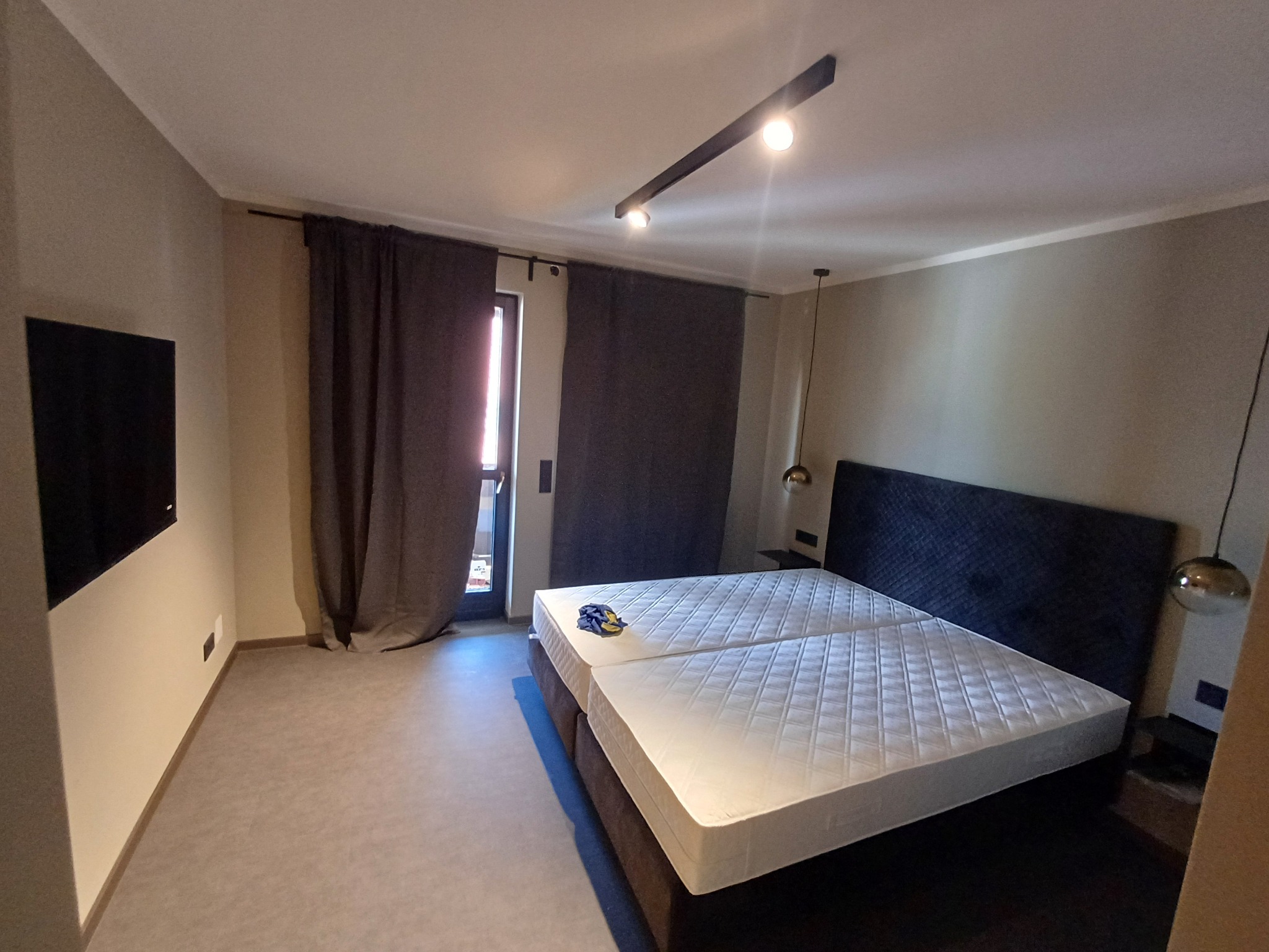 Wykończona sypialnia w Nürnberg z dużym łóżkiem, ciemnymi zasłonami i nowoczesnym oświetleniem sufitowym. Widok na balkon.