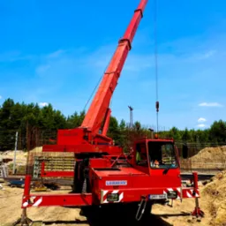 Czerwony dźwig Liebherr na placu budowy z widocznymi elementami konstrukcyjnymi i błękitnym niebem w tle, z wysuniętymi podporami stabilizującymi.