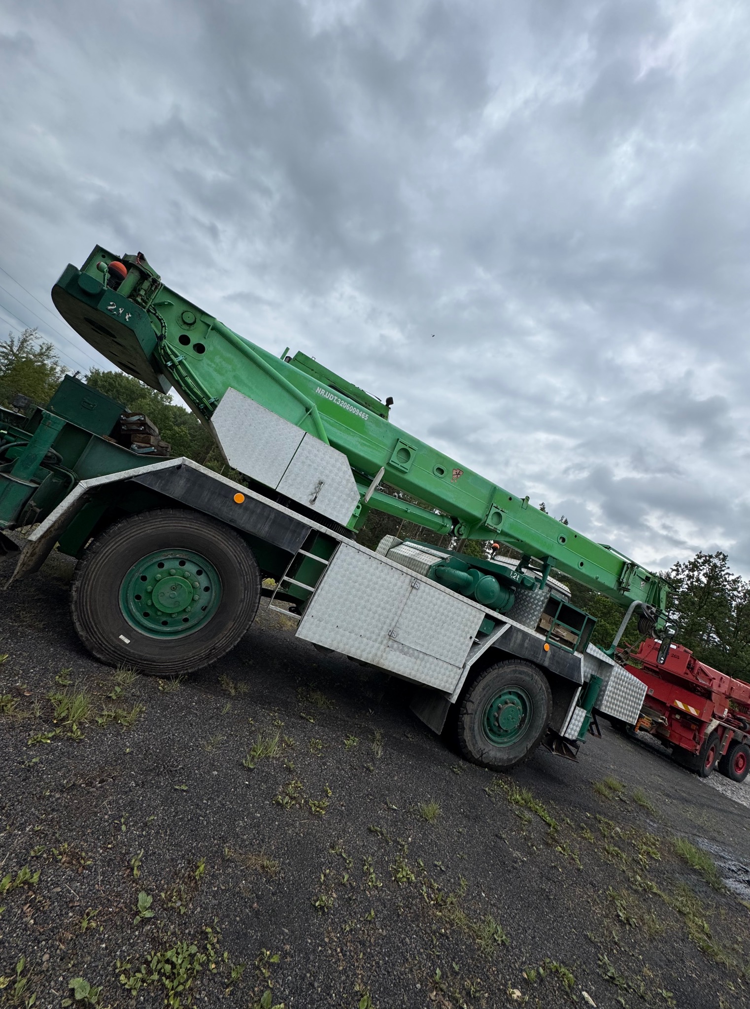 Zielony dźwig samochodowy na żwirowym podłożu, z widocznym wysięgnikiem i fragmentem drugiego, czerwonego dźwigu w tle, pod pochmurnym niebem.