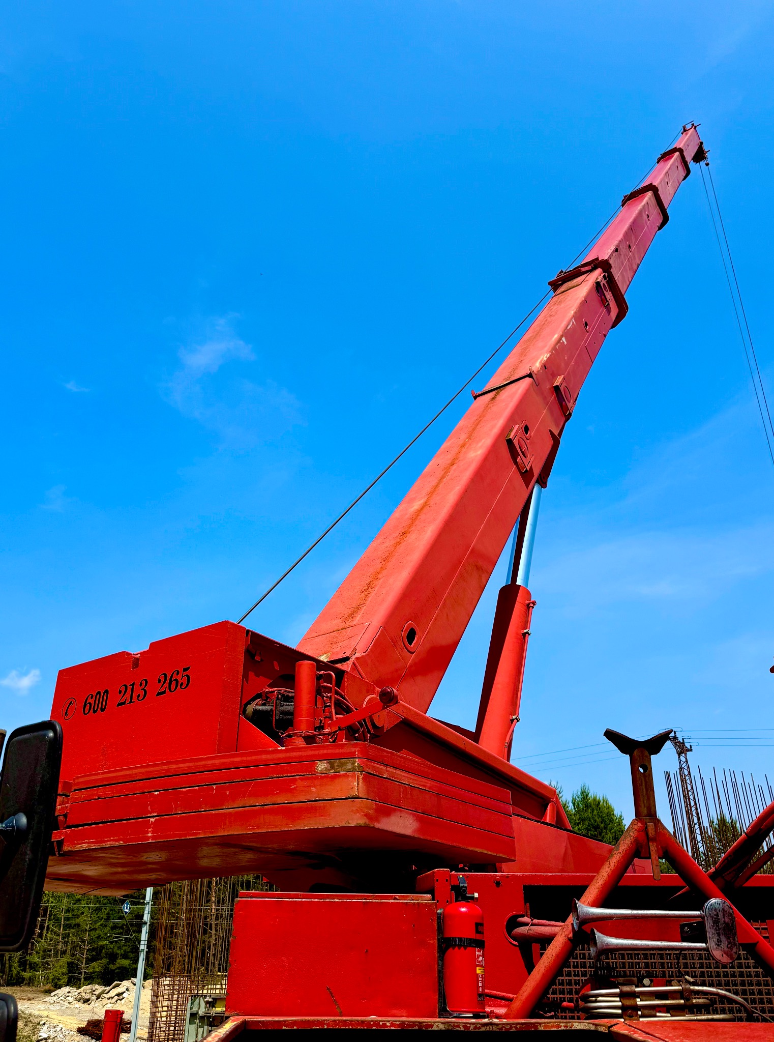 Czerwony dźwig teleskopowy na tle błękitnego nieba, widok z dołu, z numerem telefonu na obudowie. Na dole widoczne zbrojenia i drzewa.