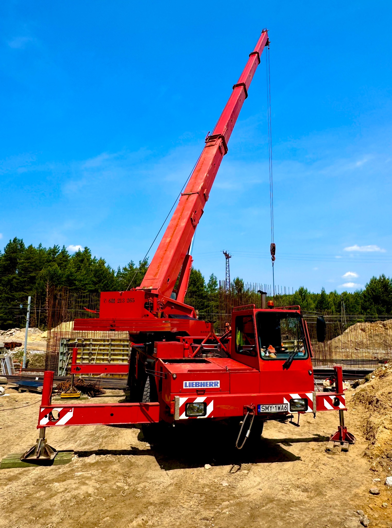 Czerwony dźwig Liebherr na placu budowy z widocznymi elementami konstrukcyjnymi i błękitnym niebem w tle, z wysuniętymi podporami stabilizującymi.
