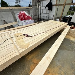 Dachy- Sylwester Niewola - Wykonywanie elementów więźby dachowej z jasnego drewna, widoczne wióry po obróbce. W tle niezadaszony budynek w trakcie budowy.