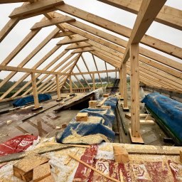 Dachy- Sylwester Niewola - Wznoszenie drewnianej więźby dachowej z widocznymi elementami konstrukcyjnymi i deskami. Na powierzchni porozrzucane wióry i fragmenty drewna. W tle widok na zewnątrz.