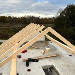 Dachy- Sylwester Niewola - Wznoszenie drewnianej więźby dachowej na betonowej płycie, widok z góry, w tle zalesiony teren. Elementy mocujące Klimas widoczne na dachu.