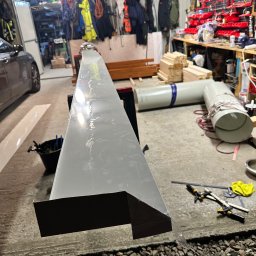 Dachy- Sylwester Niewola - Szara, metalowa obróbka blacharska z widocznym zagięciem, leżąca na warsztatowym stole w garażu, narzędzia w tle. Obróbka przygotowana do montażu.