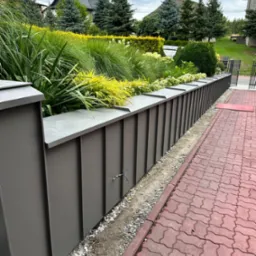 Nowoczesny, szary płot z donicami na rośliny wzdłuż czerwonej kostki brukowej. Widoczne zielone krzewy i drzewa w tle.