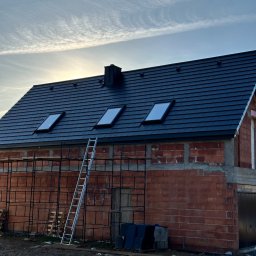 Dachy- Sylwester Niewola - Dom w trakcie budowy z czarnym dachem i trzema oknami dachowymi. Na tle błękitne niebo z delikatnymi chmurami. Przy ścianie budynku stoi drabina.