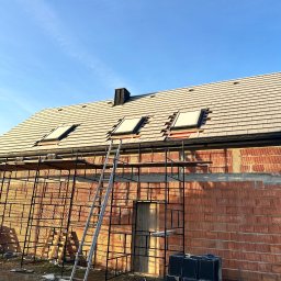 Dachy- Sylwester Niewola - Montaż okien dachowych na nowym dachu z dachówki ceramicznej. Widoczne rusztowania i materiały budowlane. Prace dekarskie w trakcie realizacji.