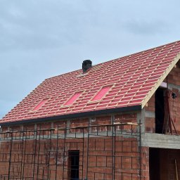 Dachy- Sylwester Niewola - Budowa dachu z czerwonej dachówki na domu z cegły. Widoczne rusztowania i komin. Dach w trakcie krycia, z widoczną membraną i łatami. Ujęcie z dołu, pochmurne niebo.