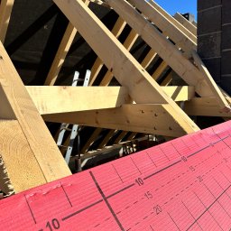 Dachy- Sylwester Niewola - Wiązar dachowy z jasnego drewna, widoczny na tle czarnej membrany i błękitnego nieba. Na pierwszym planie czerwona membrana z nadrukowaną skalą pomiarową.