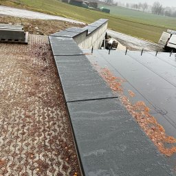 Dachy- Sylwester Niewola - Nowoczesny, płaski dach z szarej blachy na budynku w trakcie budowy, widoczne elementy mocujące i paleta z materiałami budowlanymi w tle.