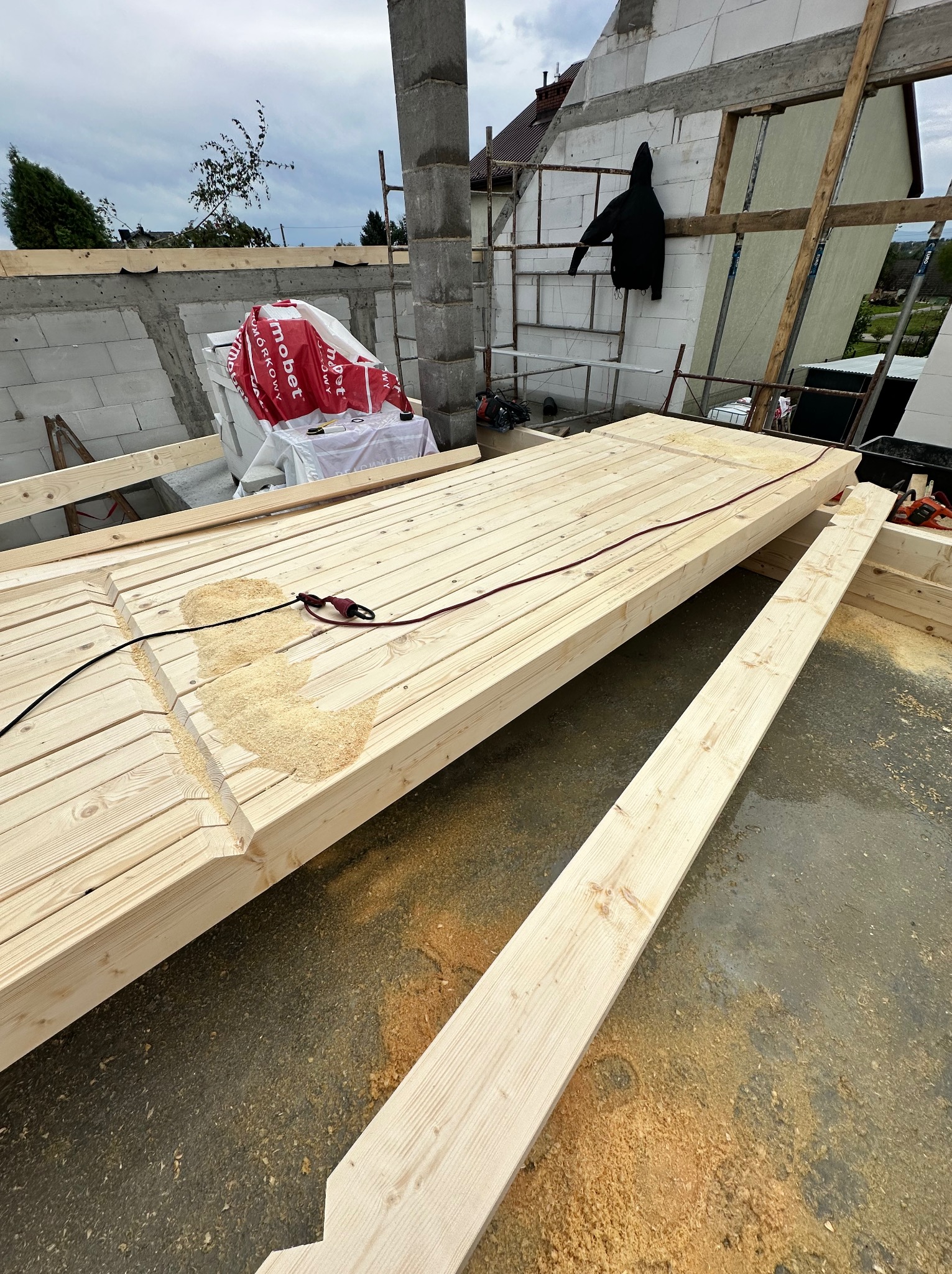 Wykonywanie elementów więźby dachowej z jasnego drewna, widoczne wióry po obróbce. W tle niezadaszony budynek w trakcie budowy.