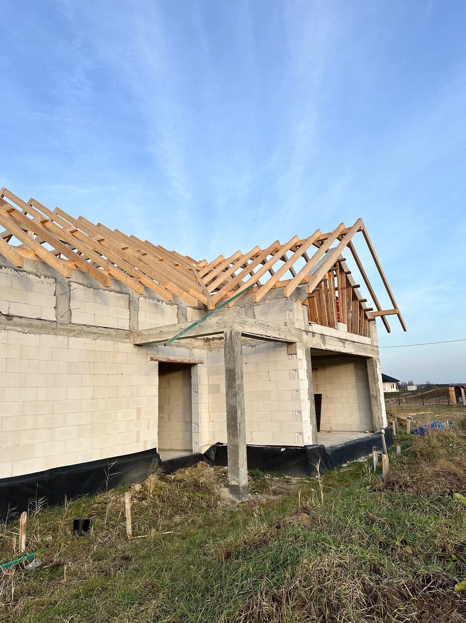 Surowy stan domu z białego bloczka z widoczną drewnianą konstrukcją dachu na tle błękitnego nieba. Widoczne detale konstrukcyjne oraz otoczenie budynku.