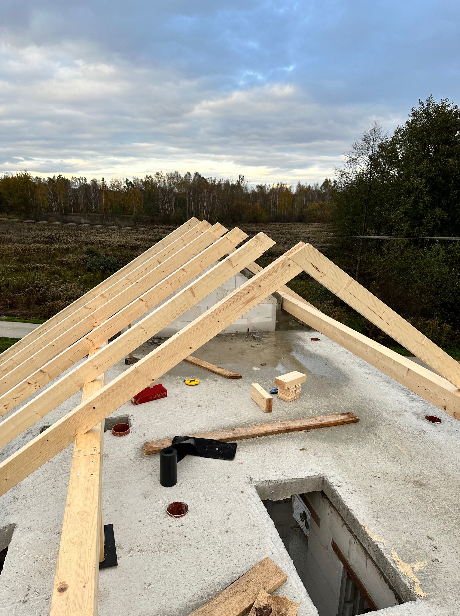 Wznoszenie drewnianej więźby dachowej na betonowej płycie, widok z góry, w tle zalesiony teren. Elementy mocujące Klimas widoczne na dachu.