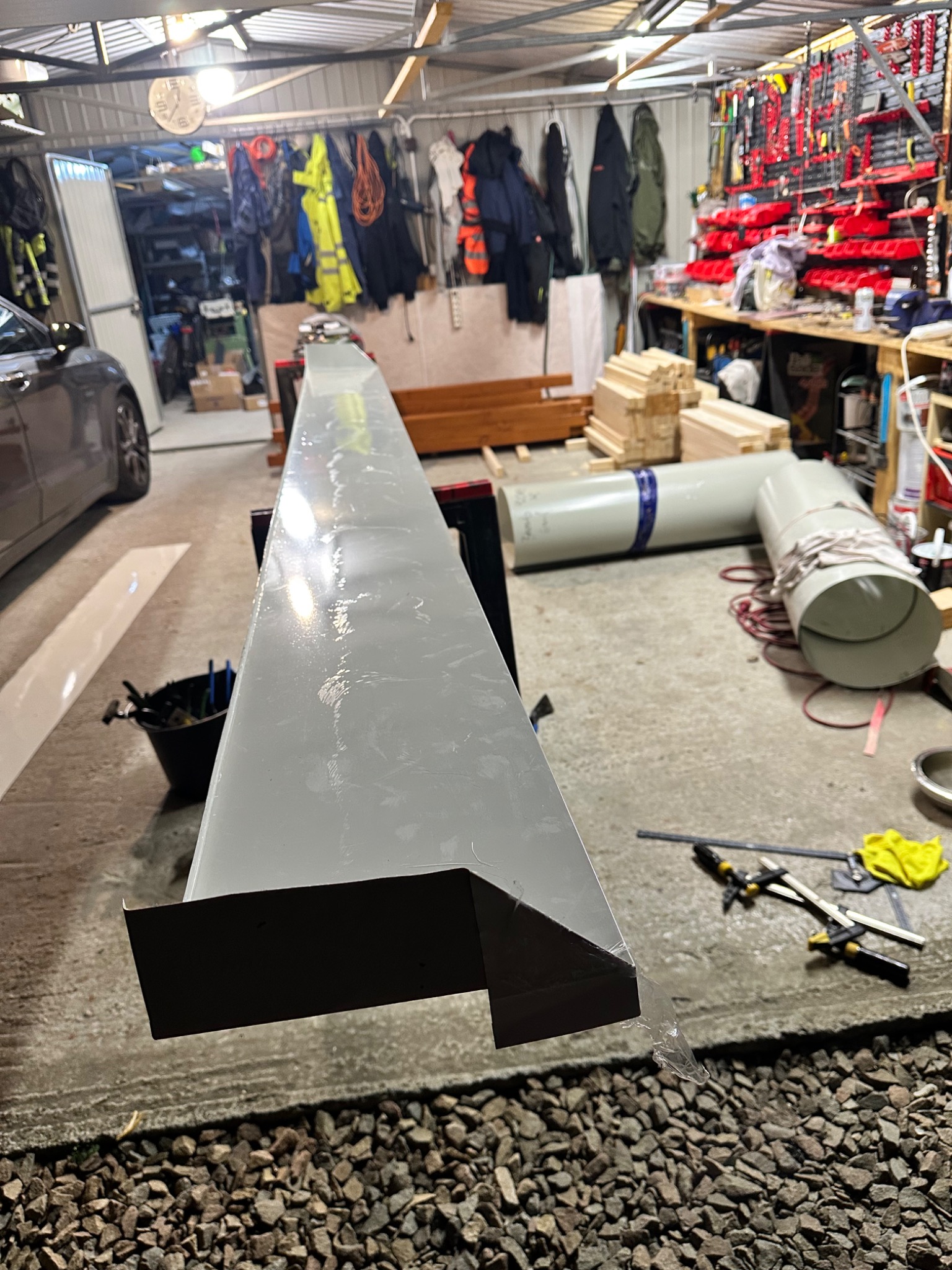 Szara, metalowa obróbka blacharska z widocznym zagięciem, leżąca na warsztatowym stole w garażu, narzędzia w tle. Obróbka przygotowana do montażu.