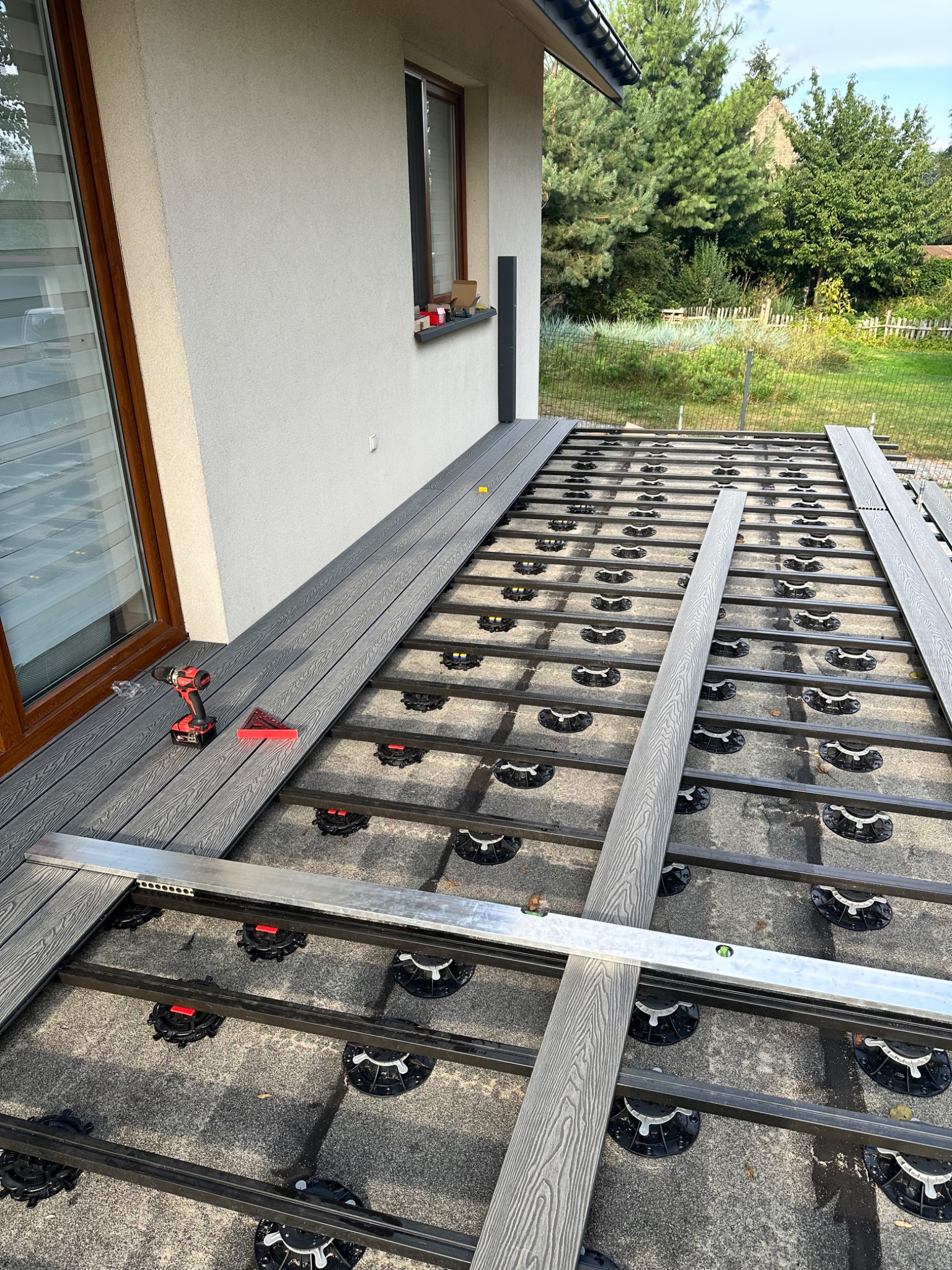 Montaż tarasu kompozytowego: widok z góry na częściowo ułożoną, szarą deskę tarasową na regulowanych wspornikach, z poziomnicą i wkrętarką obok okna.