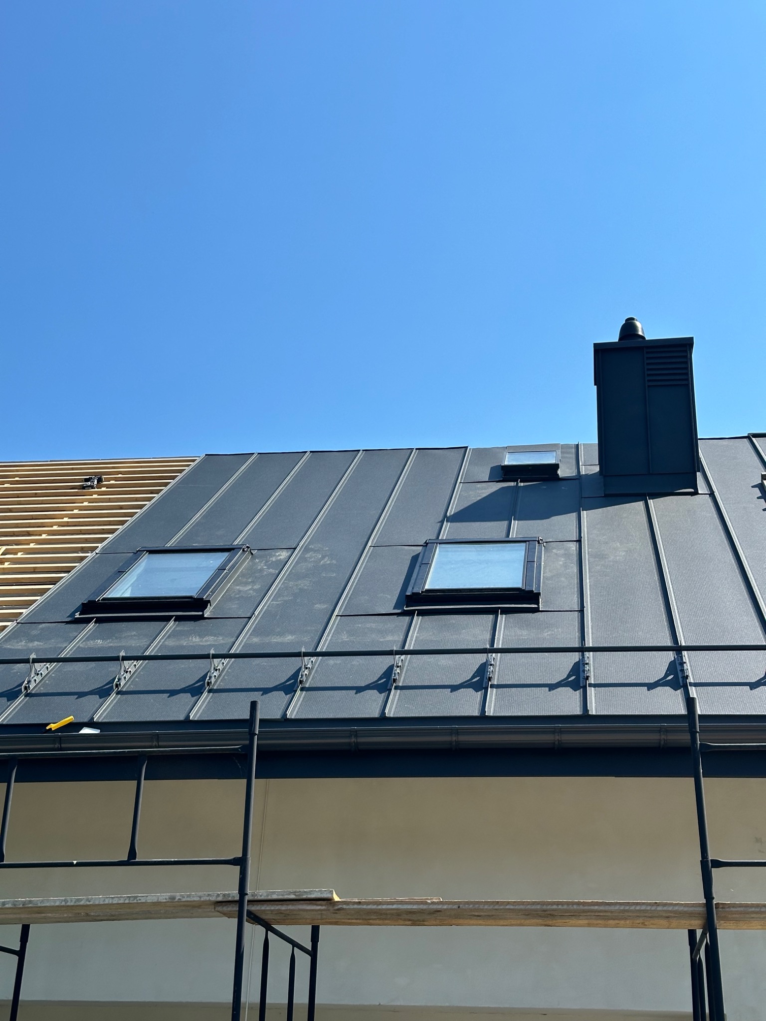 Montaż dachu z blachy na rąbek stojący z oknami dachowymi i kominem, widoczne elementy więźby dachowej, rusztowanie w dolnej części kadru, błękitne niebo.