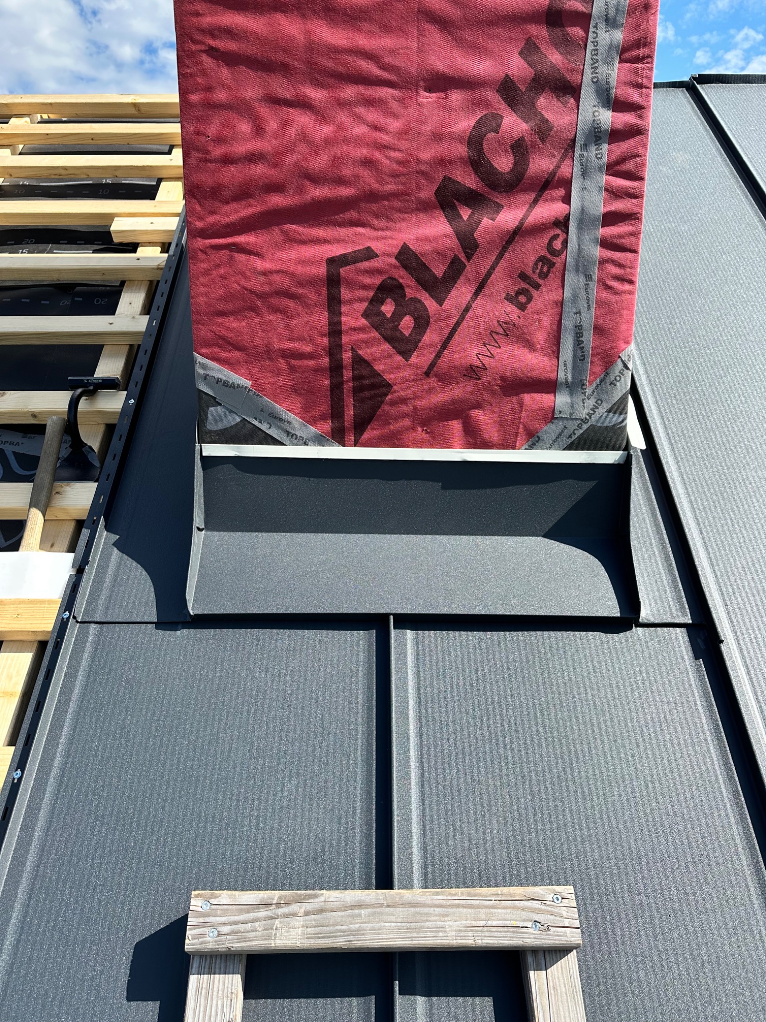 Szary dach z blachy trapezowej w trakcie montażu, widoczna membrana dachowa z czerwonym nadrukiem, fragment drewnianej konstrukcji i drabiny.