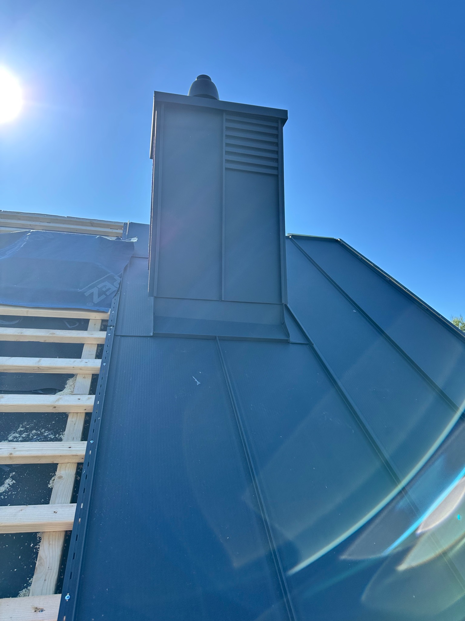 Montaż blachy na rąbek stojący na dachu z kominem w kolorze grafitowym, widok z dołu w słoneczny dzień. Częściowo widoczna więźba dachowa.