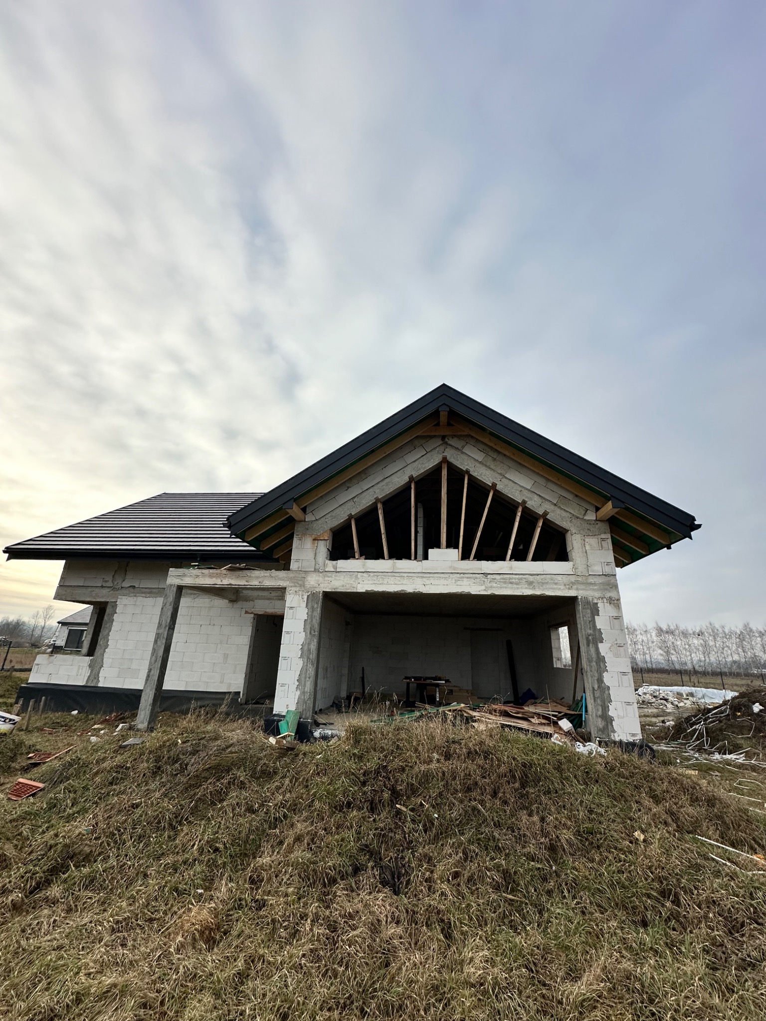 Dom w stanie surowym otwartym z częściowo wykończonym dachem z czarną dachówką, widoczne elementy więźby dachowej. Budynek z białego bloczka, otoczony trawą.