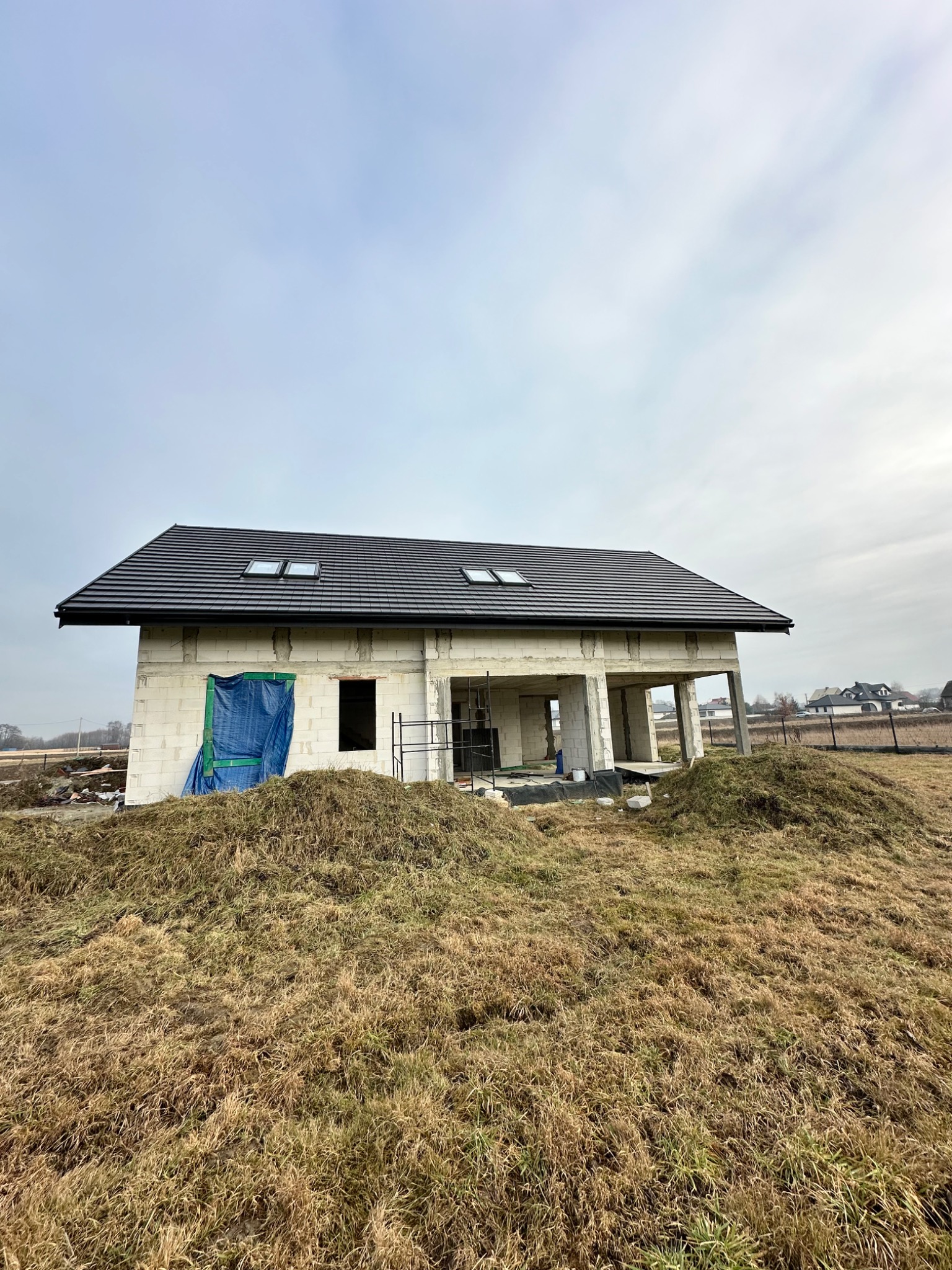 Dom w stanie surowym zamkniętym z czarną dachówką i oknami dachowymi, zabezpieczony folią, otoczony polem. Widoczne elementy konstrukcyjne i rusztowania.