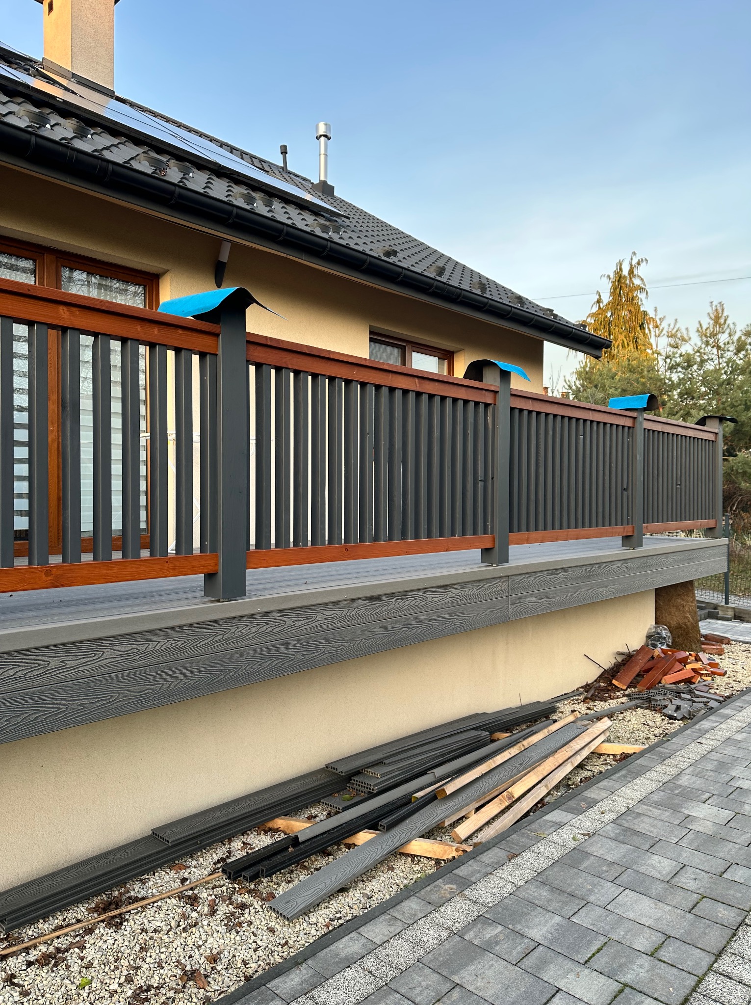 Montaż tarasu kompozytowego: widok z boku na częściowo ukończony taras z szarą deską i balustradą z drewnianymi elementami. Na ziemi leżą deski i materiały budowlane.