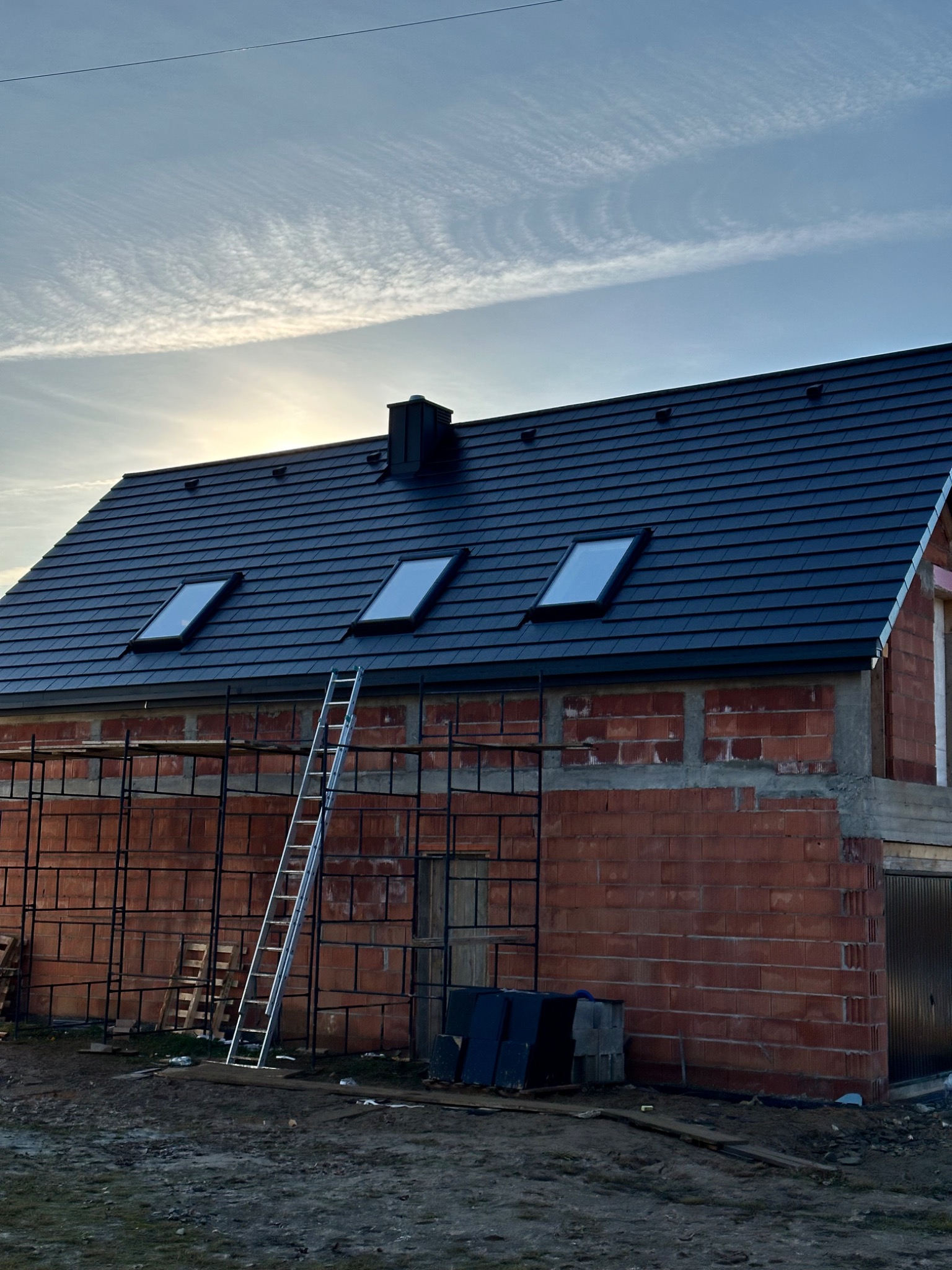 Dom w trakcie budowy z czarnym dachem i trzema oknami dachowymi. Na tle błękitne niebo z delikatnymi chmurami. Przy ścianie budynku stoi drabina.