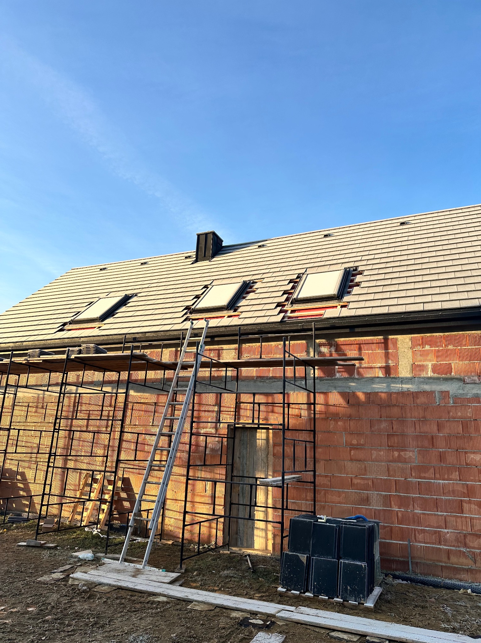 Montaż okien dachowych na nowym dachu z dachówki ceramicznej. Widoczne rusztowania i materiały budowlane. Prace dekarskie w trakcie realizacji.