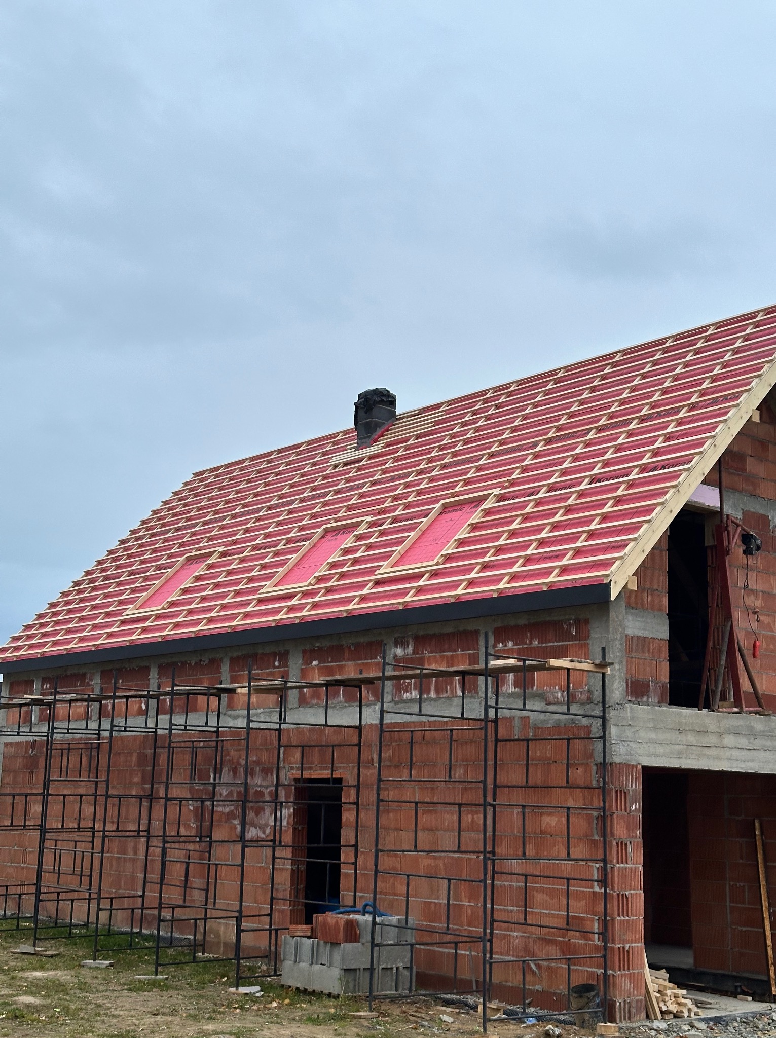 Budowa dachu z czerwonej dachówki na domu z cegły. Widoczne rusztowania i komin. Dach w trakcie krycia, z widoczną membraną i łatami. Ujęcie z dołu, pochmurne niebo.