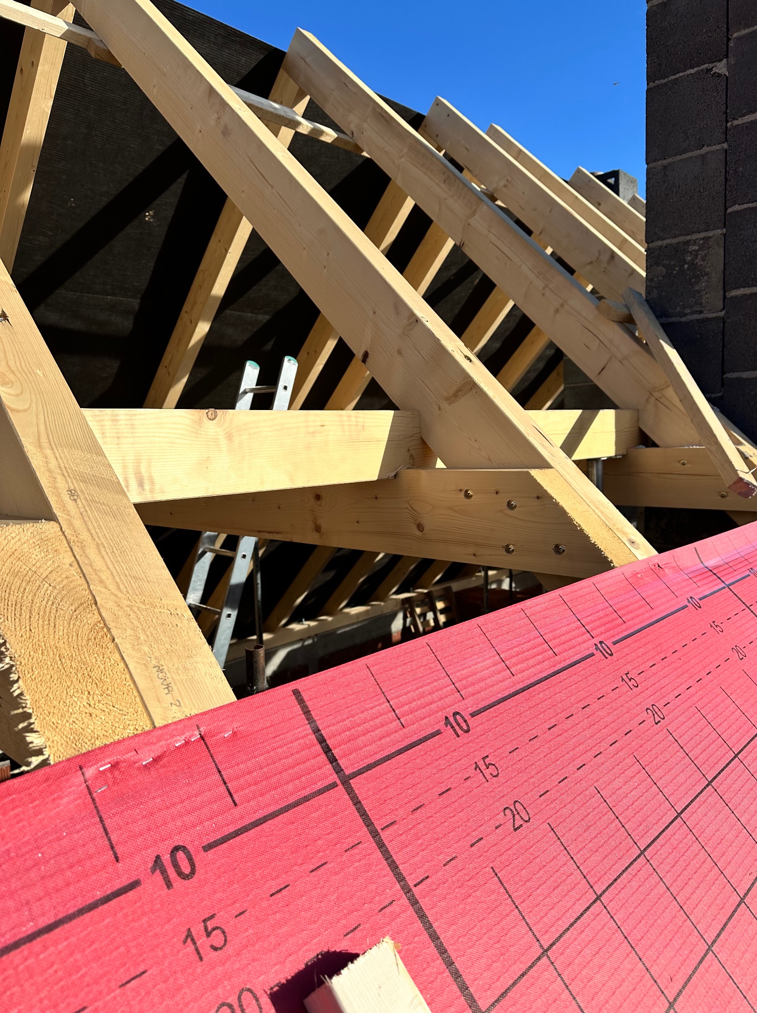 Wiązar dachowy z jasnego drewna, widoczny na tle czarnej membrany i błękitnego nieba. Na pierwszym planie czerwona membrana z nadrukowaną skalą pomiarową.