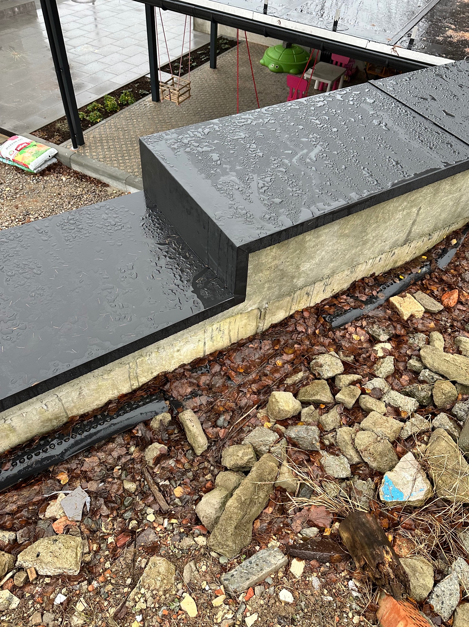 Czarny taras kompozytowy z widocznymi kroplami deszczu, betonową obudową i drenażem. W tle huśtawka i zabawki dziecięce.