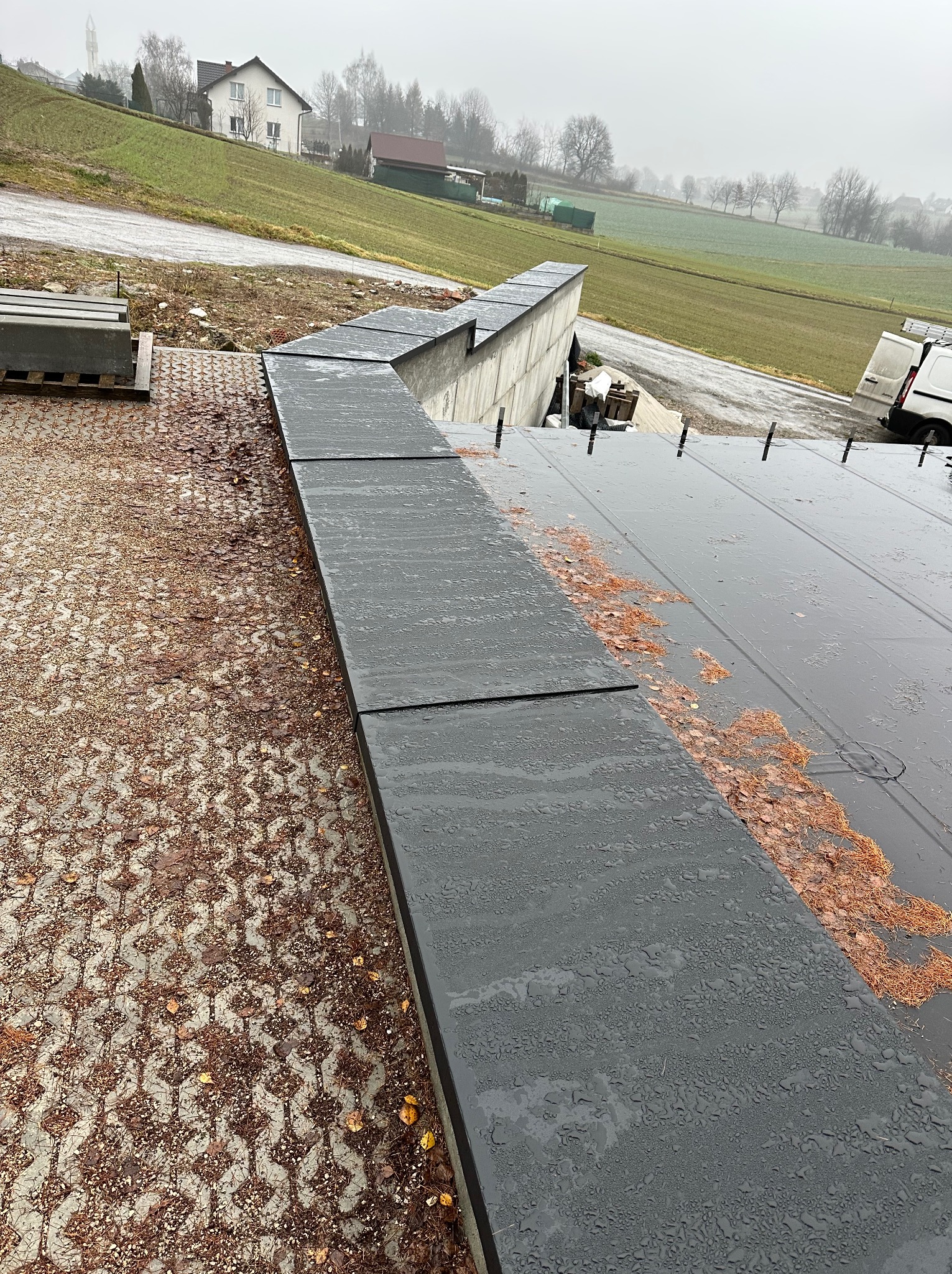 Nowoczesny, płaski dach z szarej blachy na budynku w trakcie budowy, widoczne elementy mocujące i paleta z materiałami budowlanymi w tle.