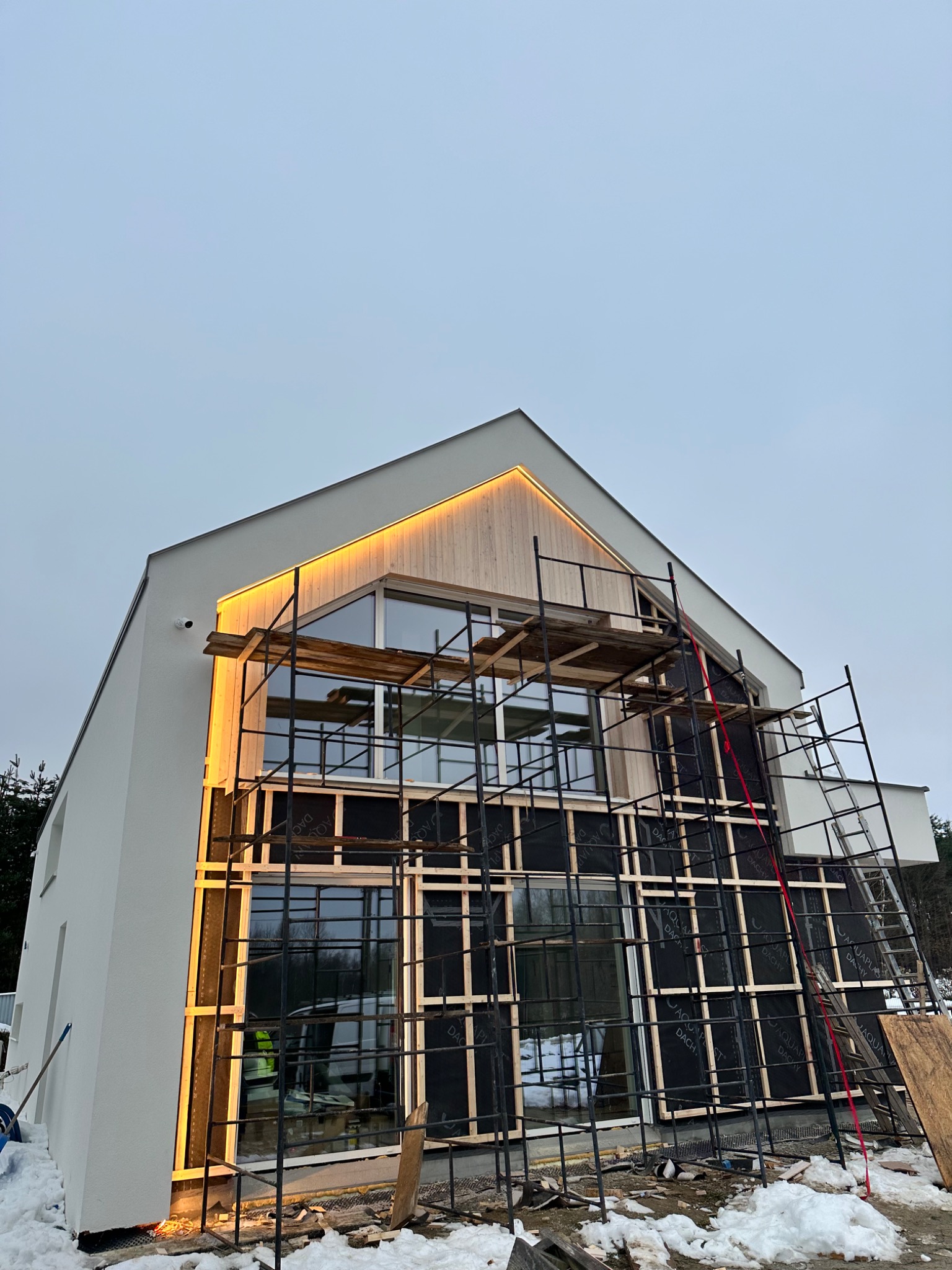 Nowoczesny dom w trakcie budowy dachu, widoczne rusztowania i oświetlenie podbitki dachowej. Ujęcie z dołu, pochmurne niebo w tle.