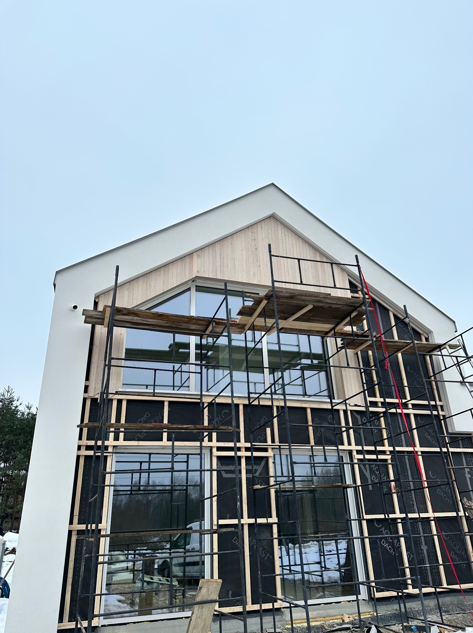 Nowoczesny dom w trakcie budowy: biała elewacja, drewniane wykończenia, okna i rusztowania. Widoczne czarne membrany dachowe z nadrukiem. Ujęcie z dołu, pochmurne niebo.