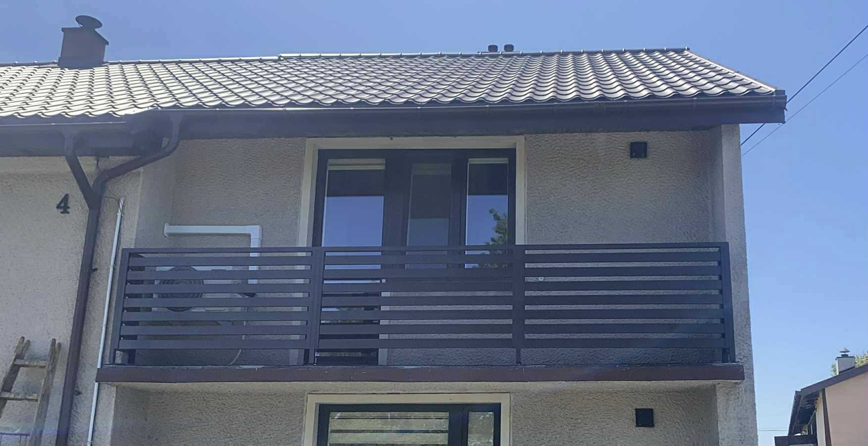 Balkon z nowoczesną, poziomą balustradą w kolorze grafitowym, zamontowaną na jasnym budynku z ciemną stolarką okienną. Widoczne detale wykończenia dachu.