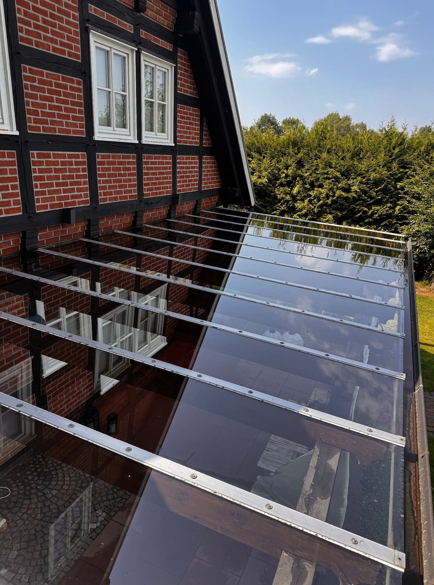 Szklany dach nad tarasem z widocznym odbiciem okien domu z muru pruskiego. Czyste powierzchnie, błękitne niebo w odbiciu.