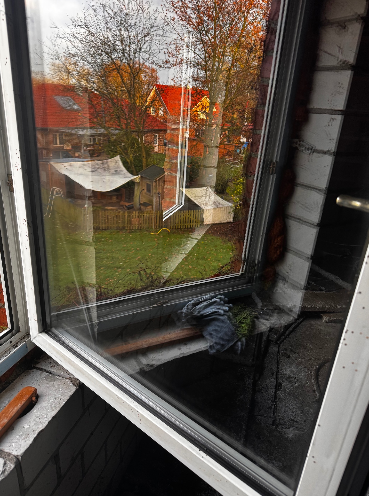 Brudne okno z widokiem na ogród, z bliska; widoczne rękawice robocze i narzędzie, co sugeruje etap przygotowania do czyszczenia.