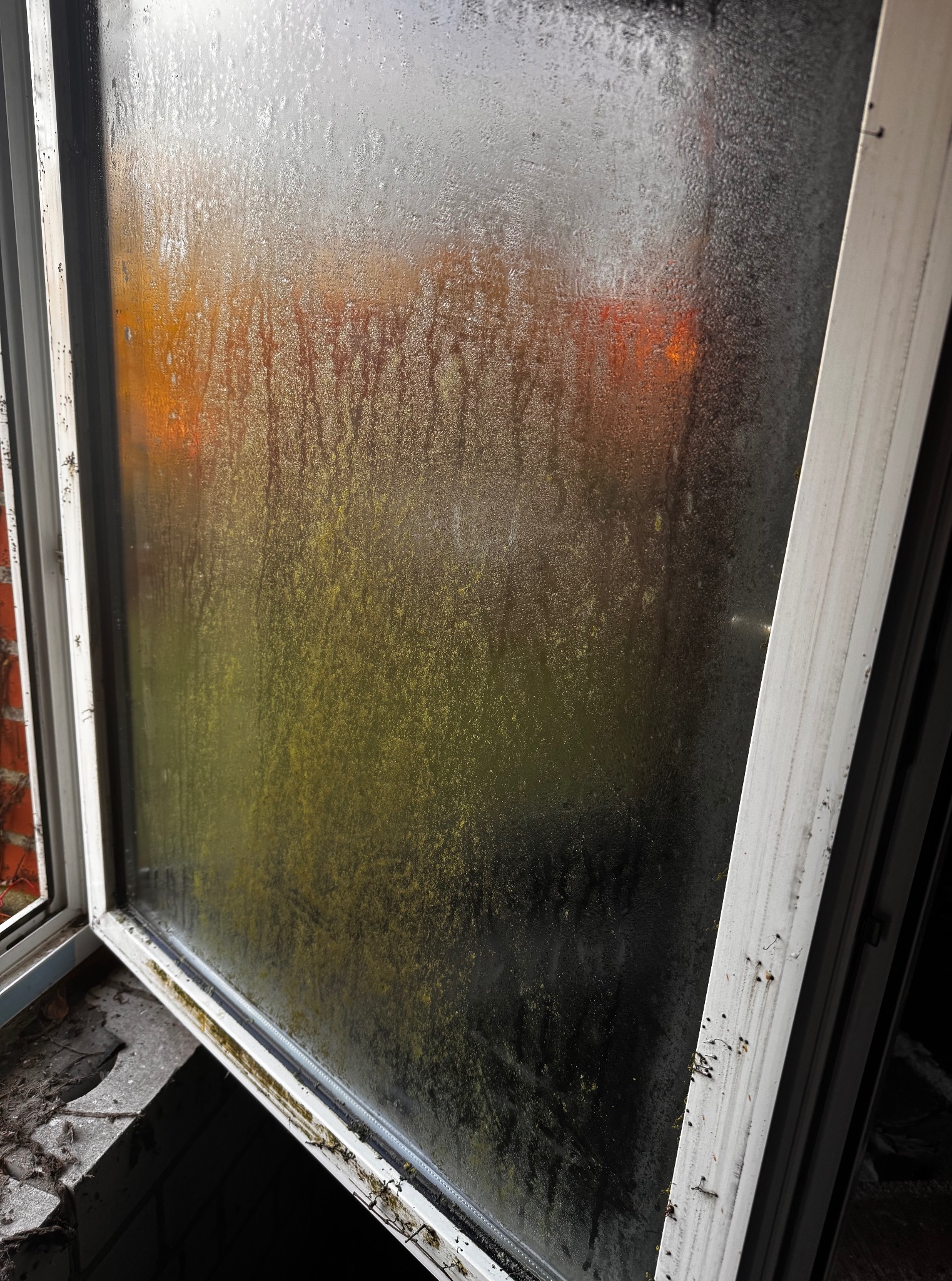 Zabrudzone okno PCV, z widocznymi zaciekami i brudem na szybie oraz ramie. Widok na zewnątrz przez brudną szybę jest nieostry.