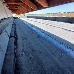 Usługi dekarskie Norbert Szczeblewski - Hydroizolacja fundamentów z widoczną warstwą izolacyjną i konstrukcją dachu w tle. Widok z perspektywy wewnętrznej na zabezpieczone fundamenty.