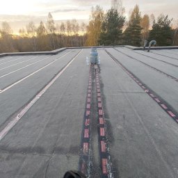 Usługi dekarskie Norbert Szczeblewski - Wykończony dach płaski z papy termozgrzewalnej, widoczne kominki wentylacyjne i butla gazowa z wodą. W tle drzewa i zachodzące słońce.