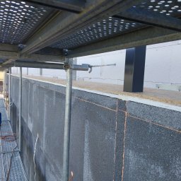 Usługi dekarskie Norbert Szczeblewski - Ocieplanie ściany zewnętrznej budynku szarym styropianem grafitowym, widoczne rusztowanie i butla gazowa. Prace izolacyjne w toku, detale montażu.