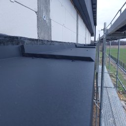 Usługi dekarskie Norbert Szczeblewski - Szczegółowe ujęcie nowej, grafitowej obróbki blacharskiej dachu z widocznymi elementami mocującymi, rusztowaniem i fragmentem ściany budynku w tle.