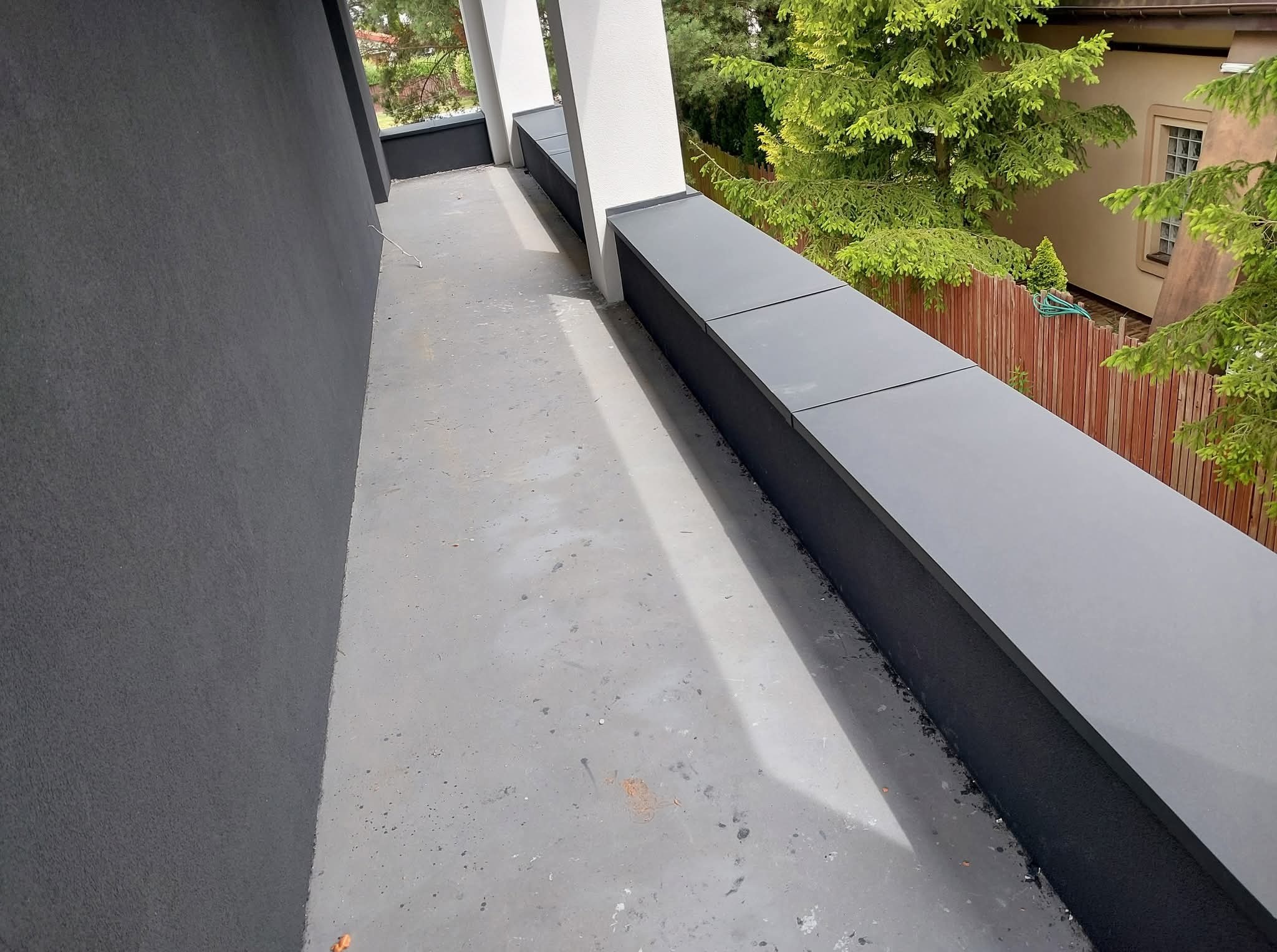 Nowoczesny, wykończony balkon z szarą balustradą i białymi kolumnami. Widok na zieleń. Minimalistyczny design, czyste linie, kątowe ujęcie.