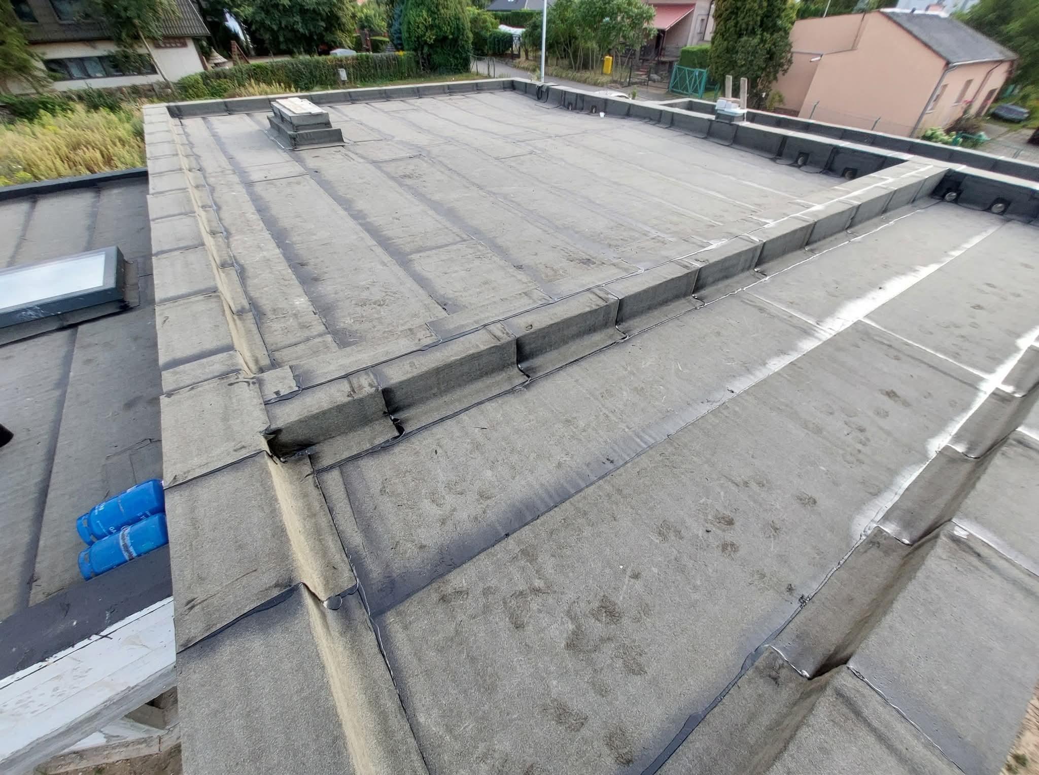 Płaski dach pokryty papą termozgrzewalną z widocznymi obróbkami blacharskimi i kominkiem wentylacyjnym. Na dachu leżą dwie butle z gazem.