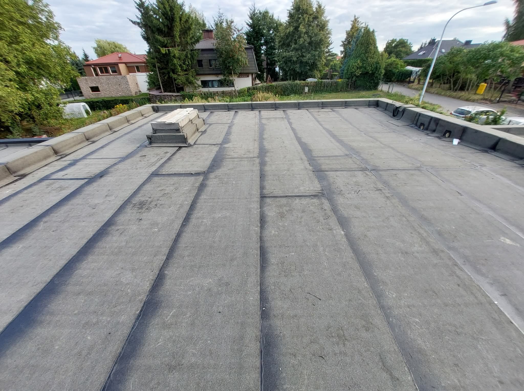 Szary dach płaski pokryty papą termozgrzewalną z kominkiem wentylacyjnym i obróbkami blacharskimi, na tle zabudowy jednorodzinnej w otoczeniu zieleni.