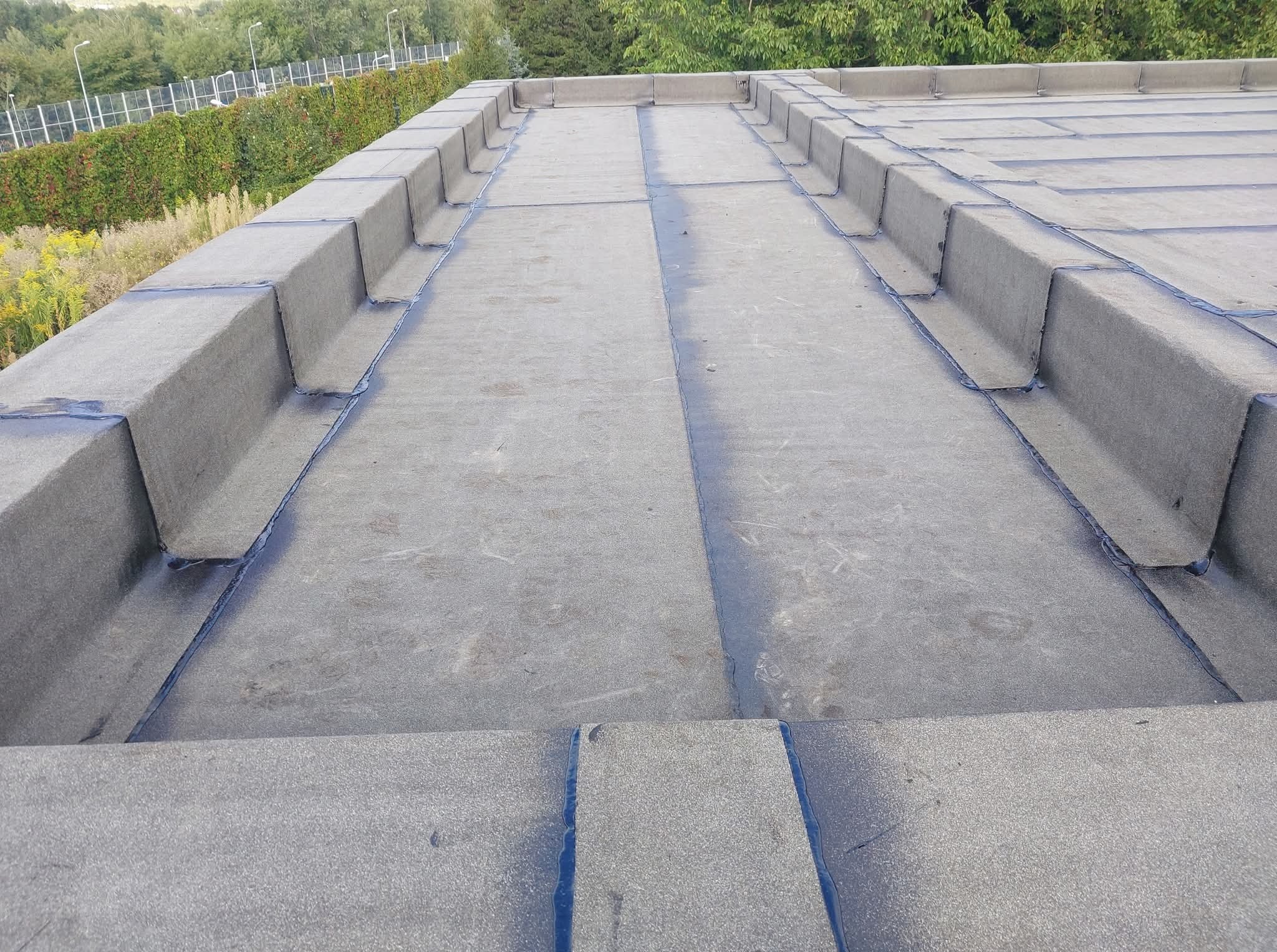 Szary, płaski dach pokryty papą termozgrzewalną z widocznymi obróbkami blacharskimi wzdłuż krawędzi i dylatacji, na tle zieleni.