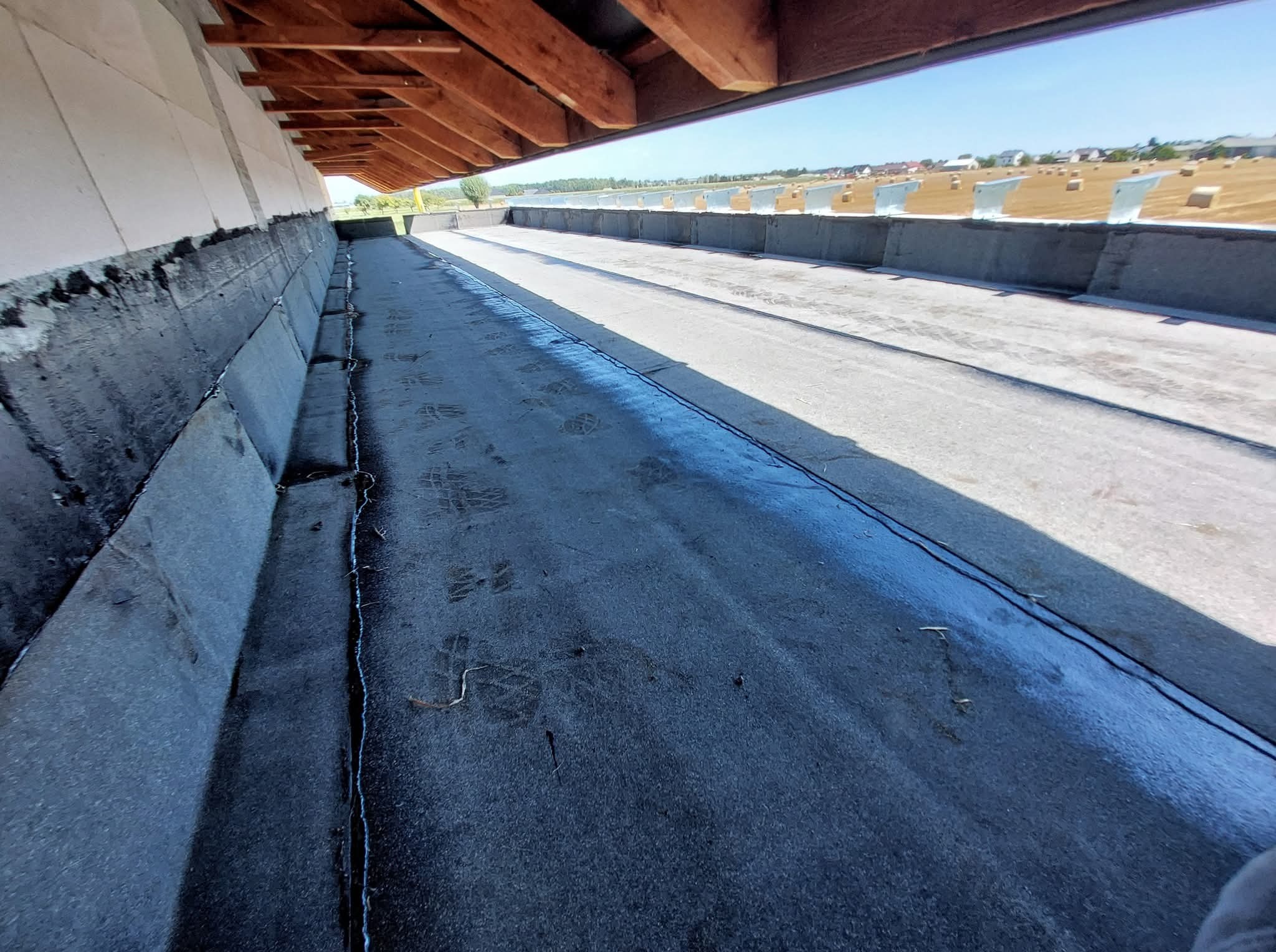 Hydroizolacja fundamentów z widoczną warstwą izolacyjną i konstrukcją dachu w tle. Widok z perspektywy wewnętrznej na zabezpieczone fundamenty.