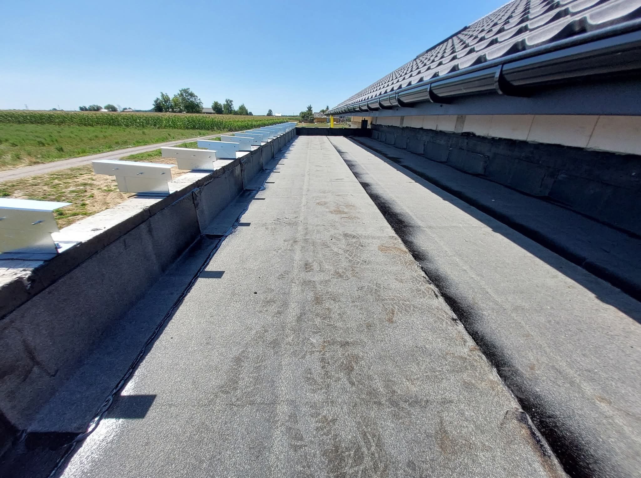 Szczegółowe ujęcie płaskiego dachu z papy termozgrzewalnej, z widocznymi obróbkami blacharskimi i zamontowanymi rynnami. W tle zieleń i błękit nieba.