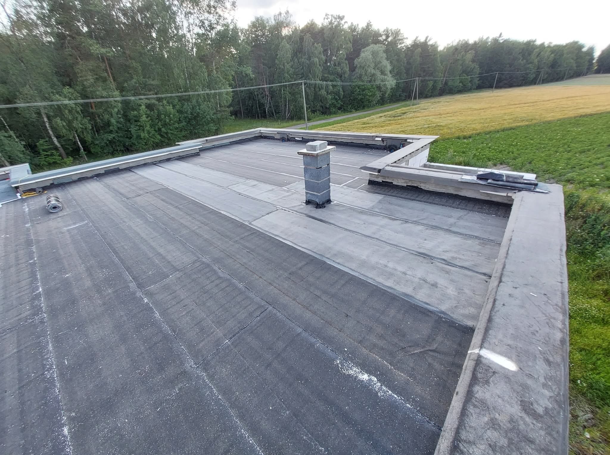 Widok z góry na płaski dach pokryty papą, z kominem z cegły szarej. W tle drzewa i pole. Widoczne obróbki blacharskie i elementy instalacji na dachu.